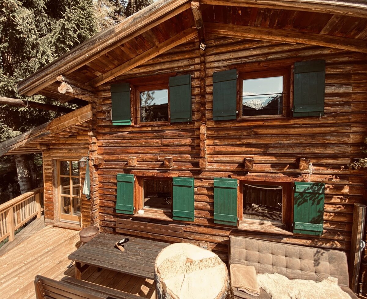 Die urige Alm aus naturbelassenem Holz mit einer einladenden Holzterrasse und grünen Fensterläden.