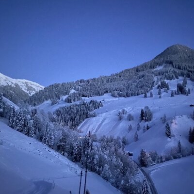 Eine malerische Winterlandschaft um die Alm mit verschneiten Berghängen und dichten Nadelwäldern unter blauem Abendhimmel.