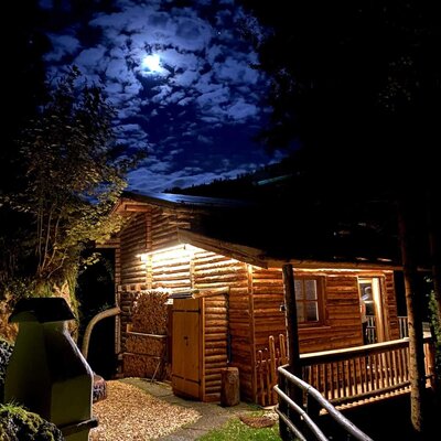 Eine einladende Alm bei Nacht, warm beleuchtet unter einem stimmungsvollen Mondscheinhimmel.