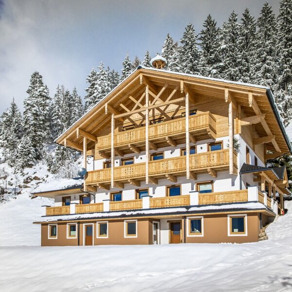 Der Bauernhof im Winter, mit traditionellen Holzbalkonen und umgeben von verschneiten Nadelbäumen.