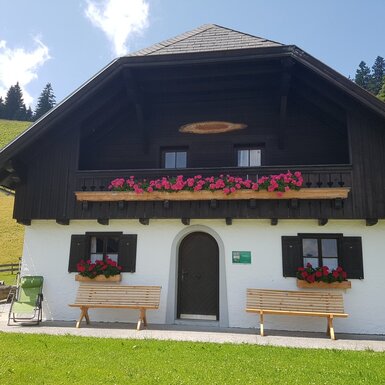 Die Almhütte mit ihrer traditionellen Fassade, einem Balkon mit Blumenkästen und Holzbänken am Eingangsbereich.
