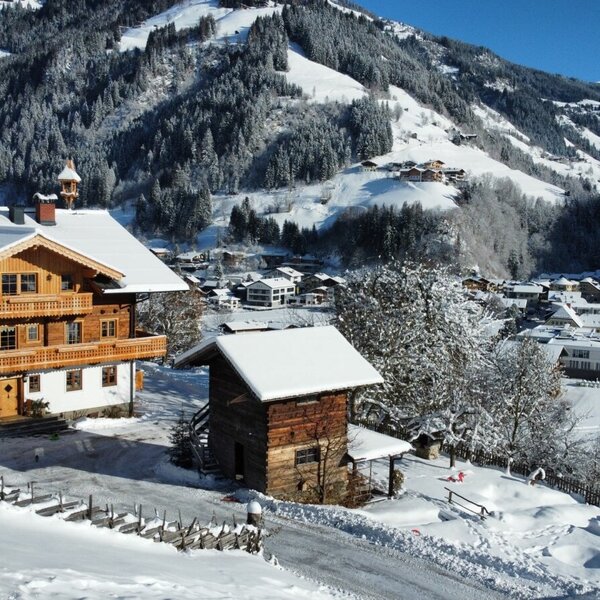 Der Bauernhof mit Holzbalkonen im Winter, umgeben von Schnee und mit Blick auf das Tal und die Berge.