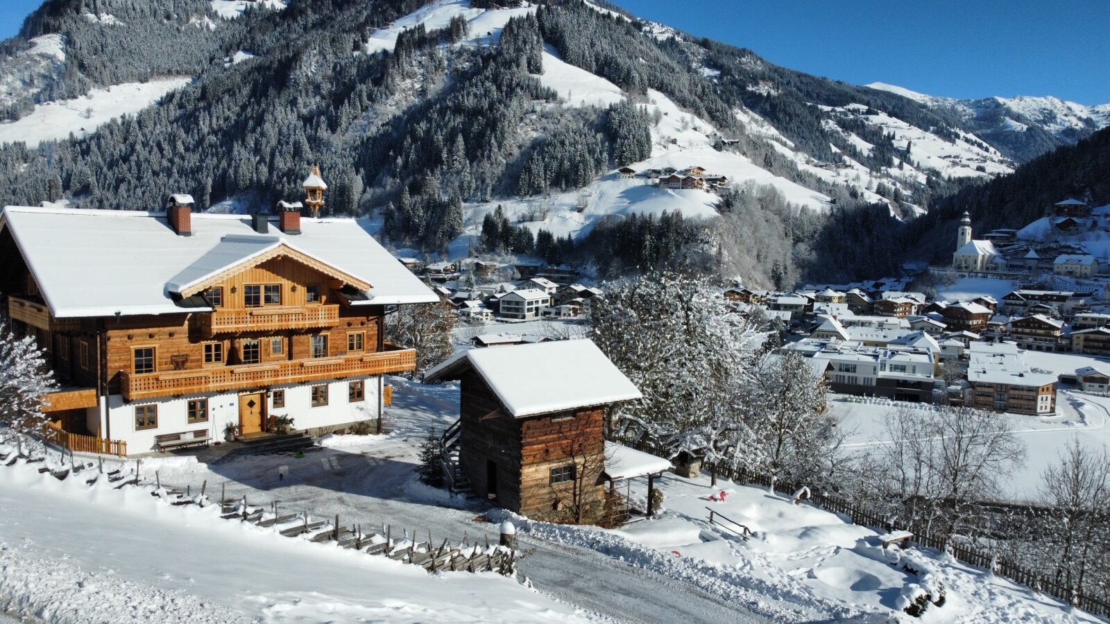 Der Bauernhof mit Holzbalkonen im Winter, umgeben von Schnee und mit Blick auf das Tal und die Berge.
