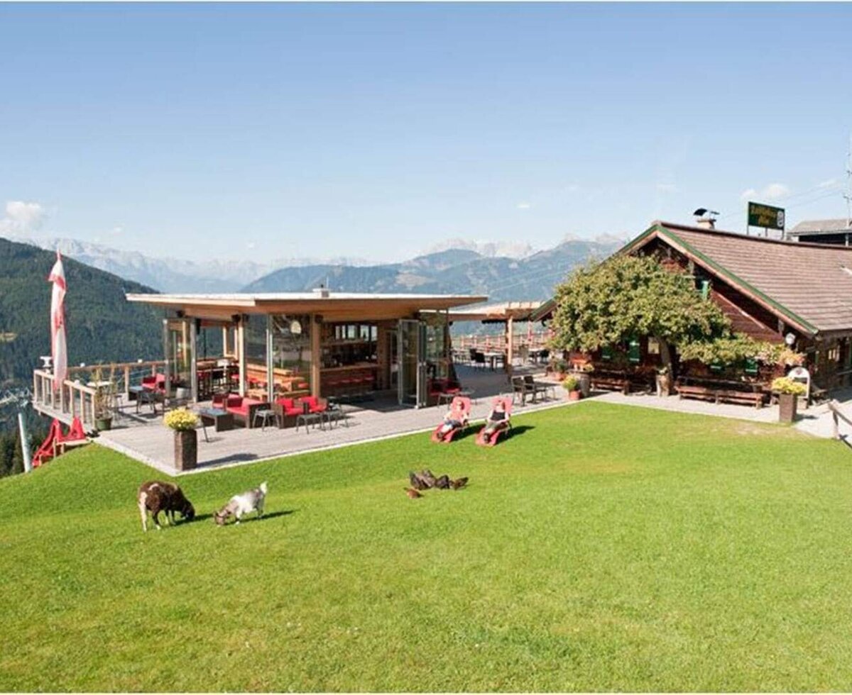 Außenansicht des Hofes mit traditionellem Gebäude und modernem Gastronomiebereich mit Terrasse, Liegewiese und Ziegen vor Bergkulisse.