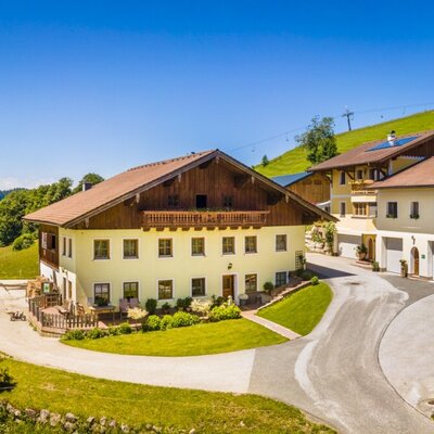 Das Anwesen des Bauernhofs mit mehreren traditionellen Gebäuden in einer grünen Berglandschaft, erschlossen durch eine Zufahrtsstraße.