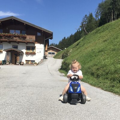 Ein Kind fährt auf einem Spielzeugtraktor vor dem Bauernhof, der kinderfreundliche Einrichtungen bietet.