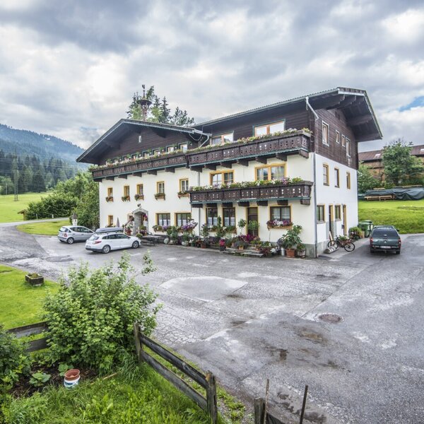 Außenansicht des Bauernhofs mit Parkplatz und Balkonen mit Blumenkästen vor einer Berglandschaft.