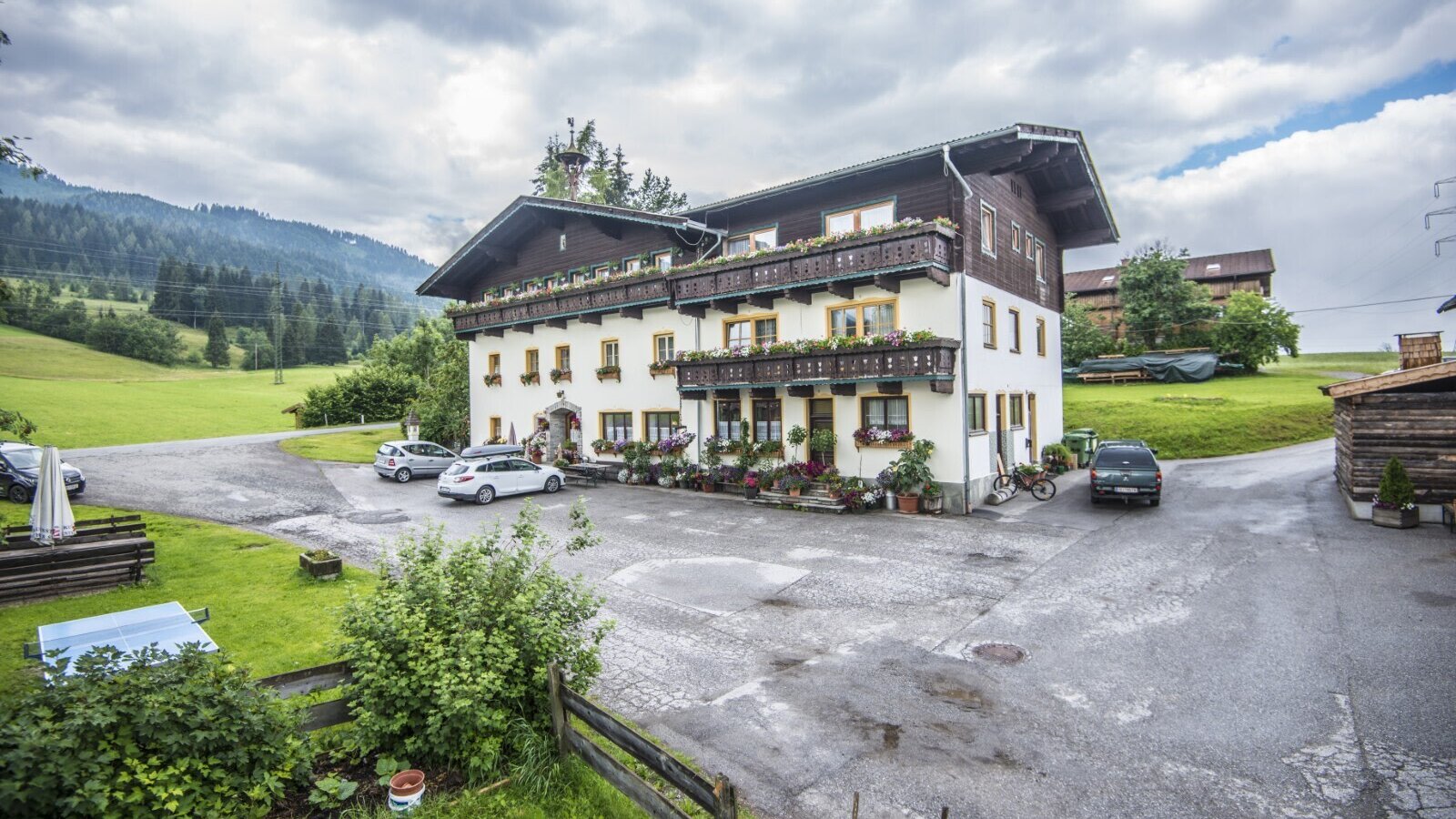 Außenansicht des Bauernhofs mit Parkplatz und Balkonen mit Blumenkästen vor einer Berglandschaft.