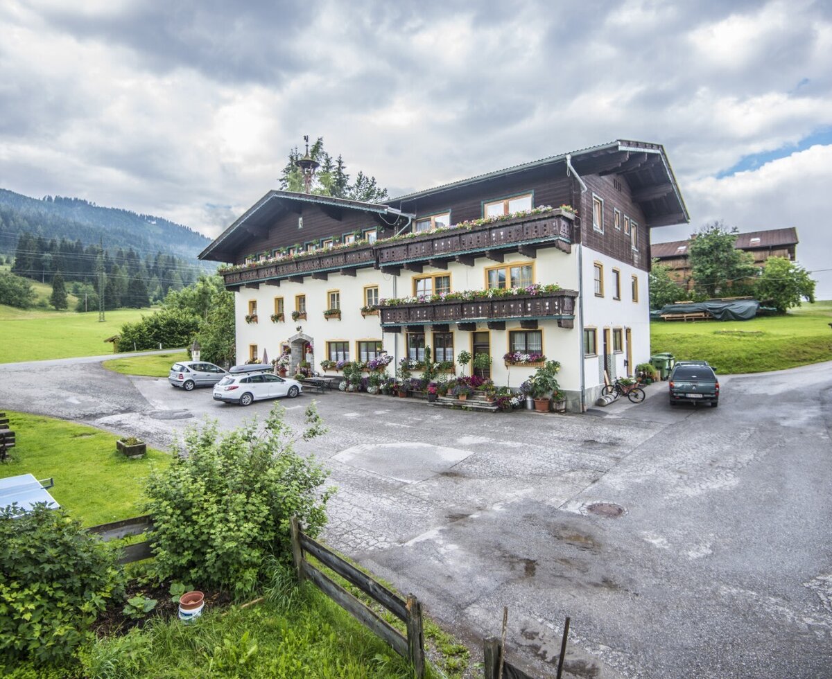 Außenansicht des Bauernhofs mit Parkplatz und Balkonen mit Blumenkästen vor einer Berglandschaft.
