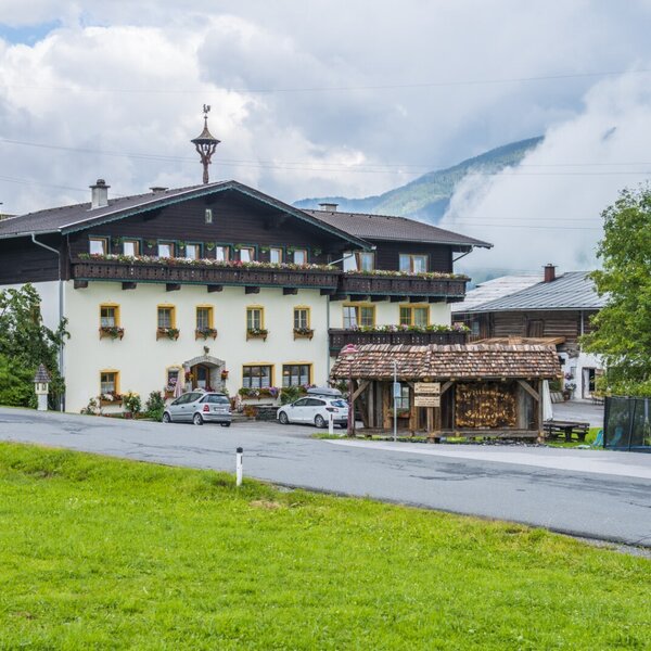 Der Bauernhof mit seiner traditionellen Fassade, Balkonen mit Blumenkästen und Parkmöglichkeiten vor der Bergkulisse.