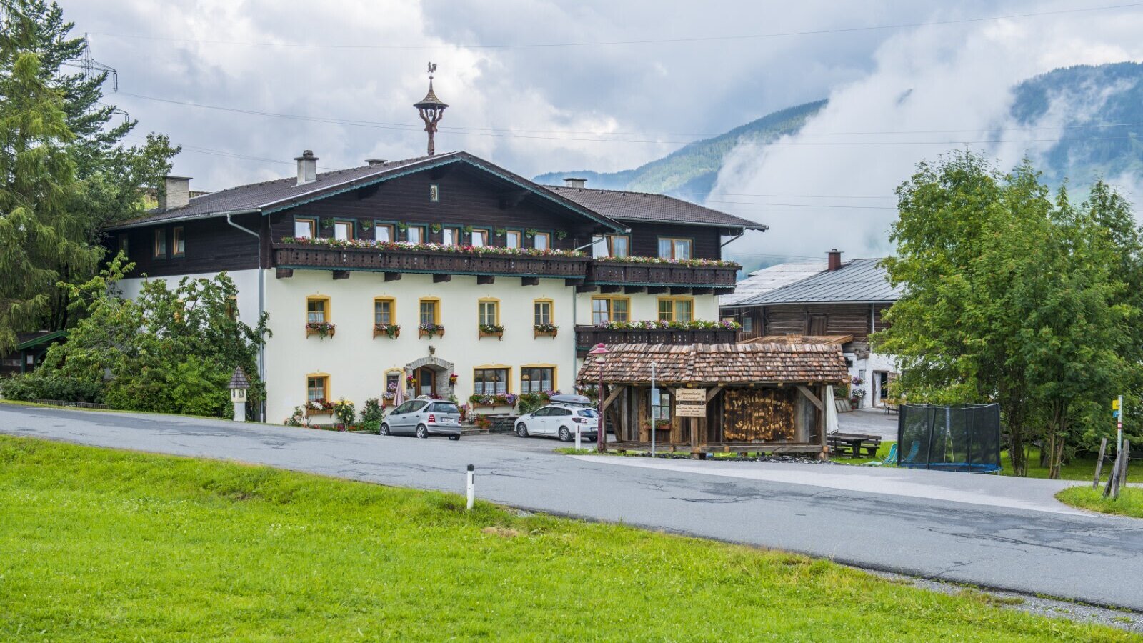 Der Bauernhof mit seiner traditionellen Fassade, Balkonen mit Blumenkästen und Parkmöglichkeiten vor der Bergkulisse.