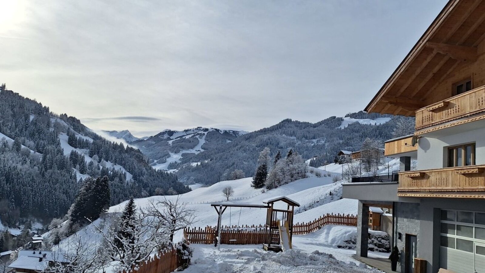 Blick vom Bauernhof auf die verschneite Berglandschaft, inklusive Kinderspielplatz und Holzbalkonen am Gebäude.