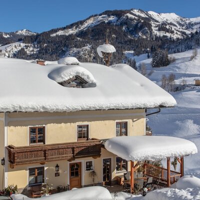 Der Bauernhof mit gelber Fassade, Holzbalkon und überdachter Terrasse, umgeben von verschneiten Bergen.