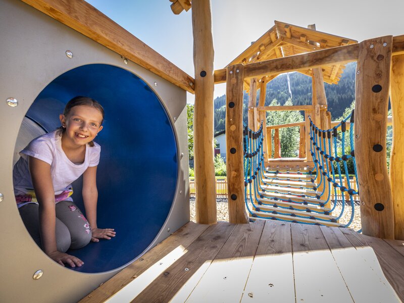 Ein Spielplatzbereich am Bauernhof mit Holzelementen, einem Tunnel und einer Seilbrücke für Kinder.