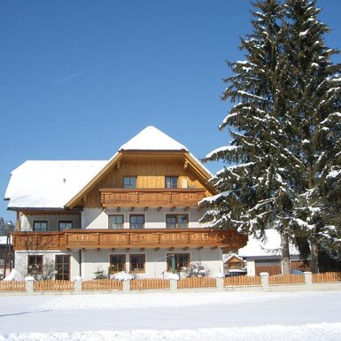 Der verschneite Bauernhof mit Holzbalkonen und einem großen Fichtenbaum.