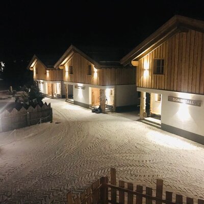 Die beleuchteten Gebäude des Bauernhofs in der Nacht, mit schneebedecktem Gelände und dem "GOAS STALL".