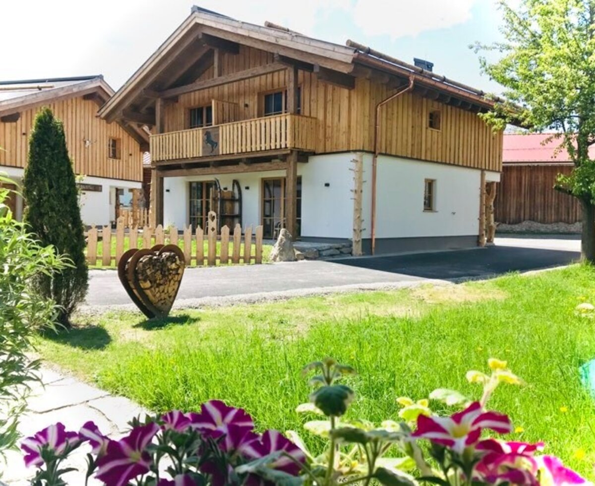 Der Bauernhof mit traditioneller Holzfassade, Balkon und dem Gartenbereich mit Blumenbeeten.