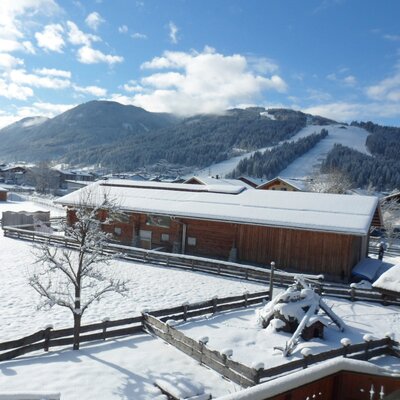 Blick vom Bauernhof auf die verschneite Winterlandschaft mit Skipiste und umliegenden Gebäuden.