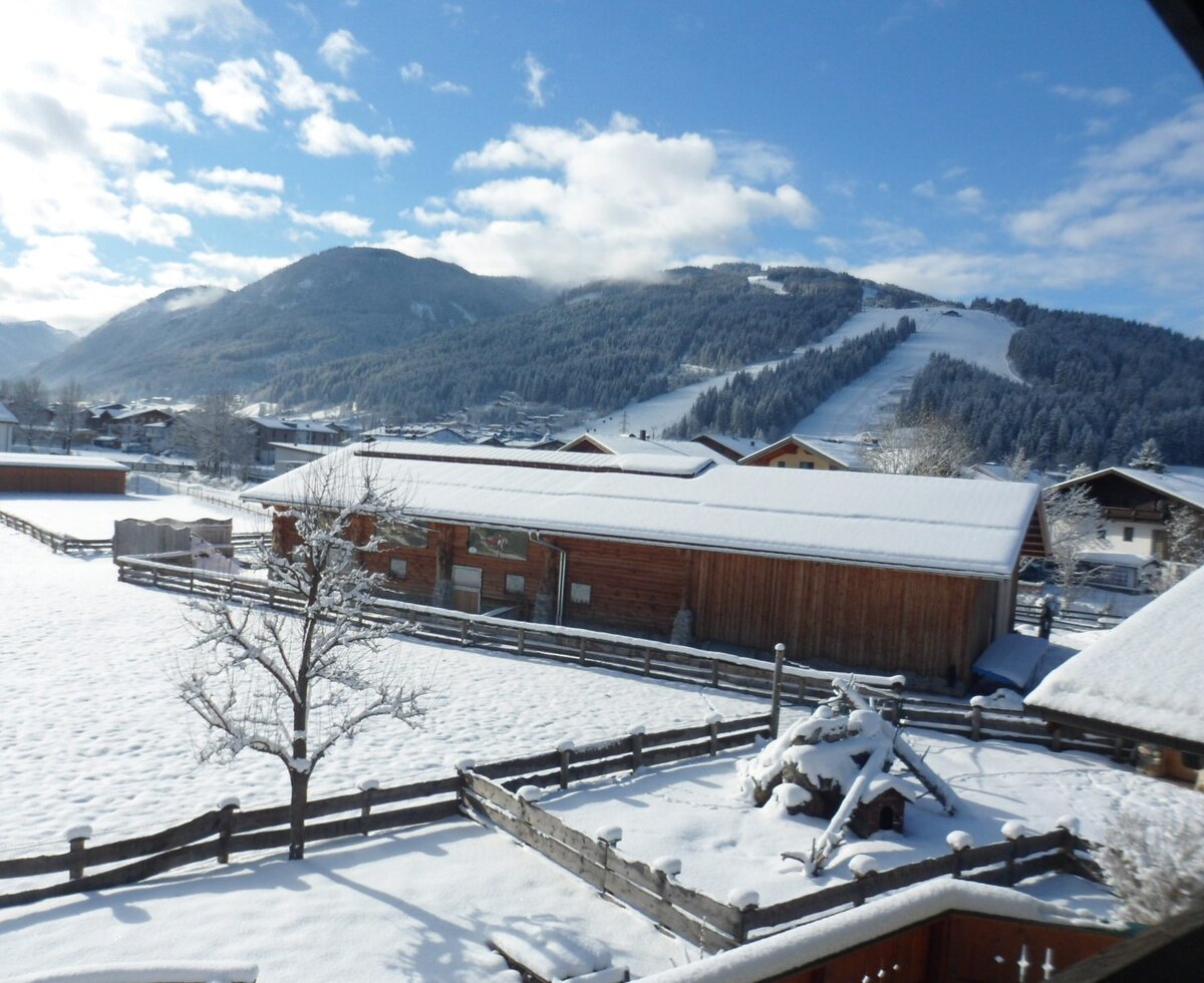 Blick vom Bauernhof auf die verschneite Winterlandschaft mit Skipiste und umliegenden Gebäuden.