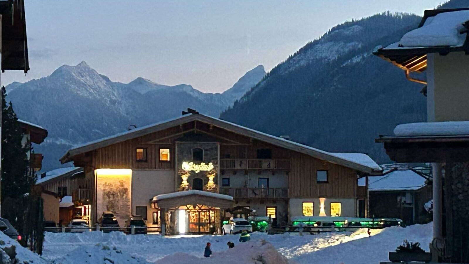 Der Bauernhof im Winter, mit beleuchtetem Eingang und schneebedeckten Bergen im Hintergrund.