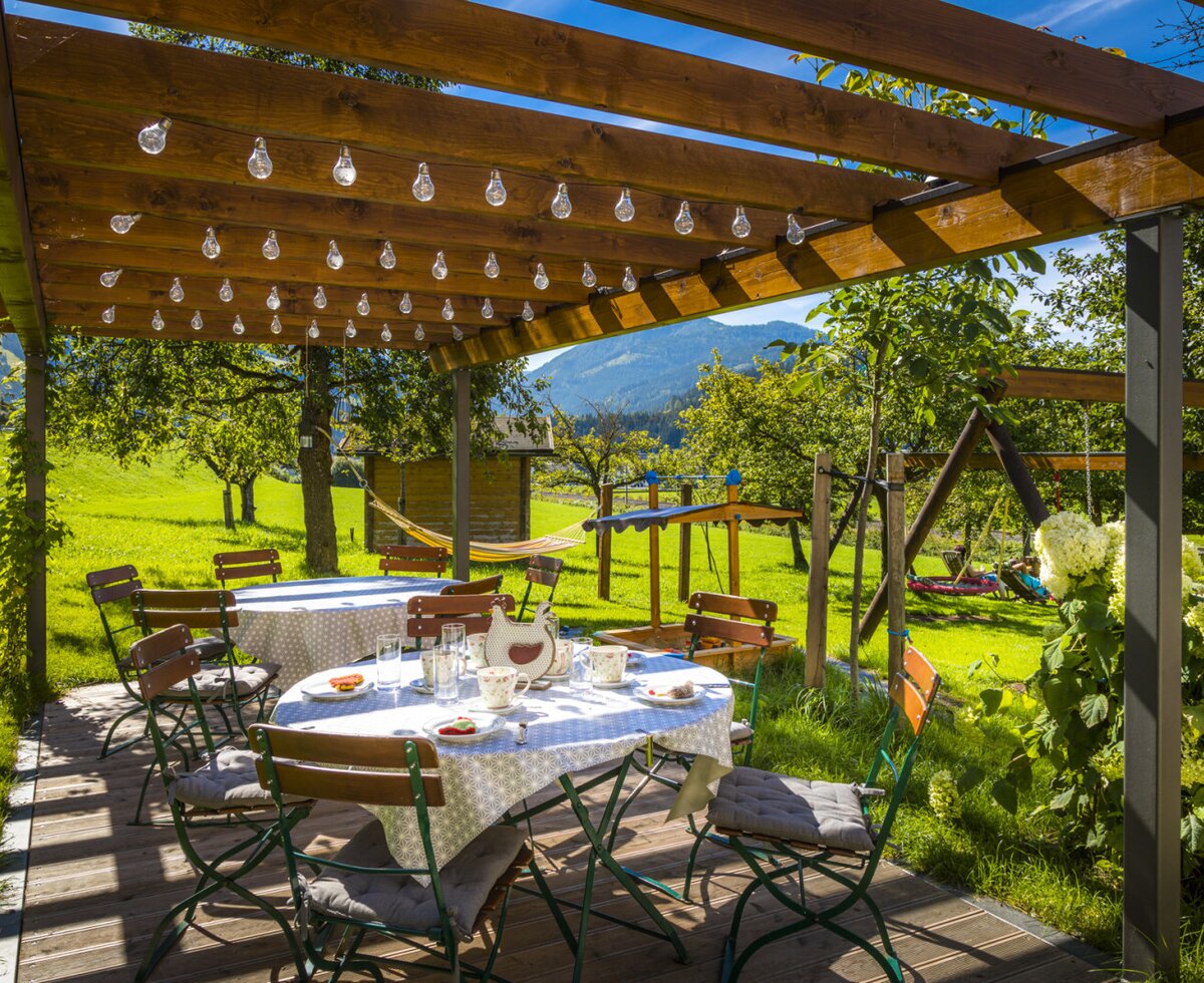 Überdachte Terrasse des Bauernhofs mit Esstischen und Blick auf den Garten mit Kinderspielplatz und die Berge.