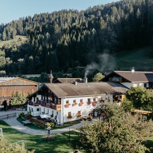 Das Äußere des Bauernhofs mit mehreren Gebäuden, darunter das Hauptgebäude mit Balkonen, einer großen Liegewiese mit Kinderspielgeräten und bewaldeten Hängen im Hintergrund.
