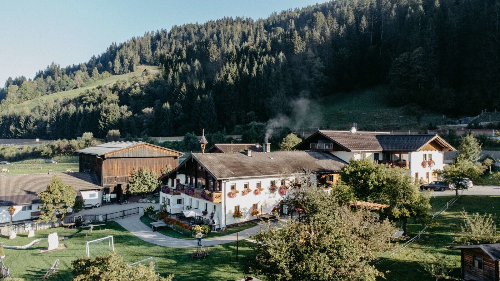 Das Äußere des Bauernhofs mit mehreren Gebäuden, darunter das Hauptgebäude mit Balkonen, einer großen Liegewiese mit Kinderspielgeräten und bewaldeten Hängen im Hintergrund.
