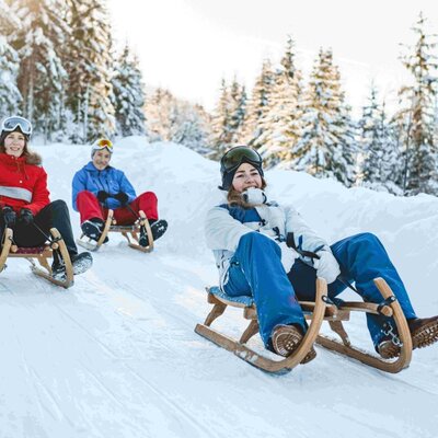 Drei Personen rodeln auf einem verschneiten Hang, eine Winteraktivität in der Umgebung des Bauernhofs.