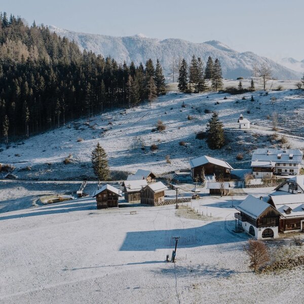 Der Bauernhof in einer verschneiten Winterlandschaft mit mehreren Gebäuden, umliegenden Wäldern und Bergen.