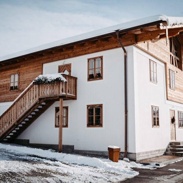 Die winterliche Außenansicht des Bauernhofs mit weißer Fassade, holzverkleideten Elementen und einer Außentreppe, die zu einem oberen Eingang führt.
