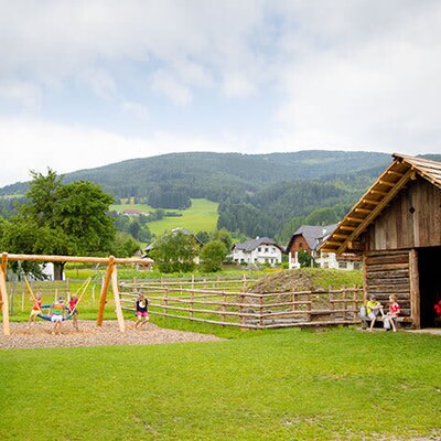 Kinderspielplatz des Bauernhofs mit Schaukeln und spielenden Kindern vor einer Holzscheune und Berglandschaft.