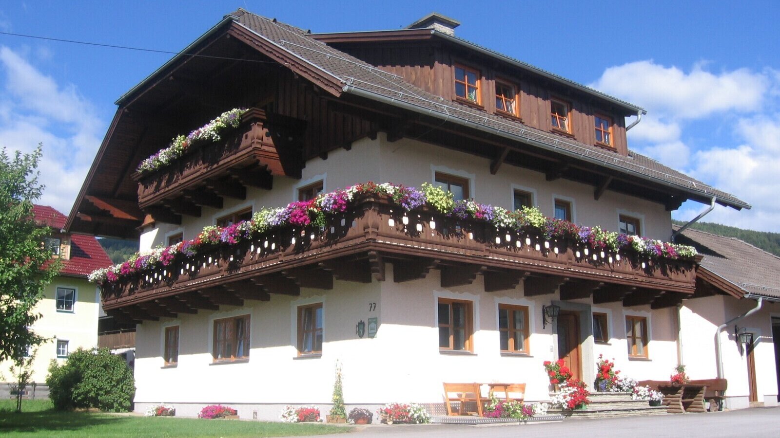 Außenansicht des Bauernhofs mit mehreren Balkonen, geschmückt mit bunten Blumenkästen, und einem Eingangsbereich mit Holzbänken.