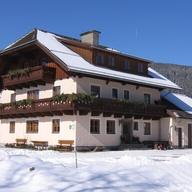 Der Bauernhof im Winter mit Holzbalkonen, verschneitem Dach und einem Berg im Hintergrund.