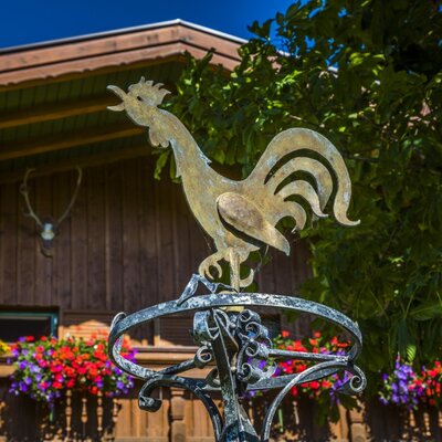 Der Metallhahn als Wetterfahne vor der Holzfassade des Bauernhofs mit bunten Blumenkästen und Hirschgeweih.