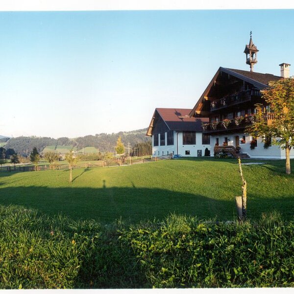 Der Bauernhof mit Glockenturm, umgeben von grünen Wiesen und bewaldeten Hügeln, inklusive Schaukel auf dem Gelände.