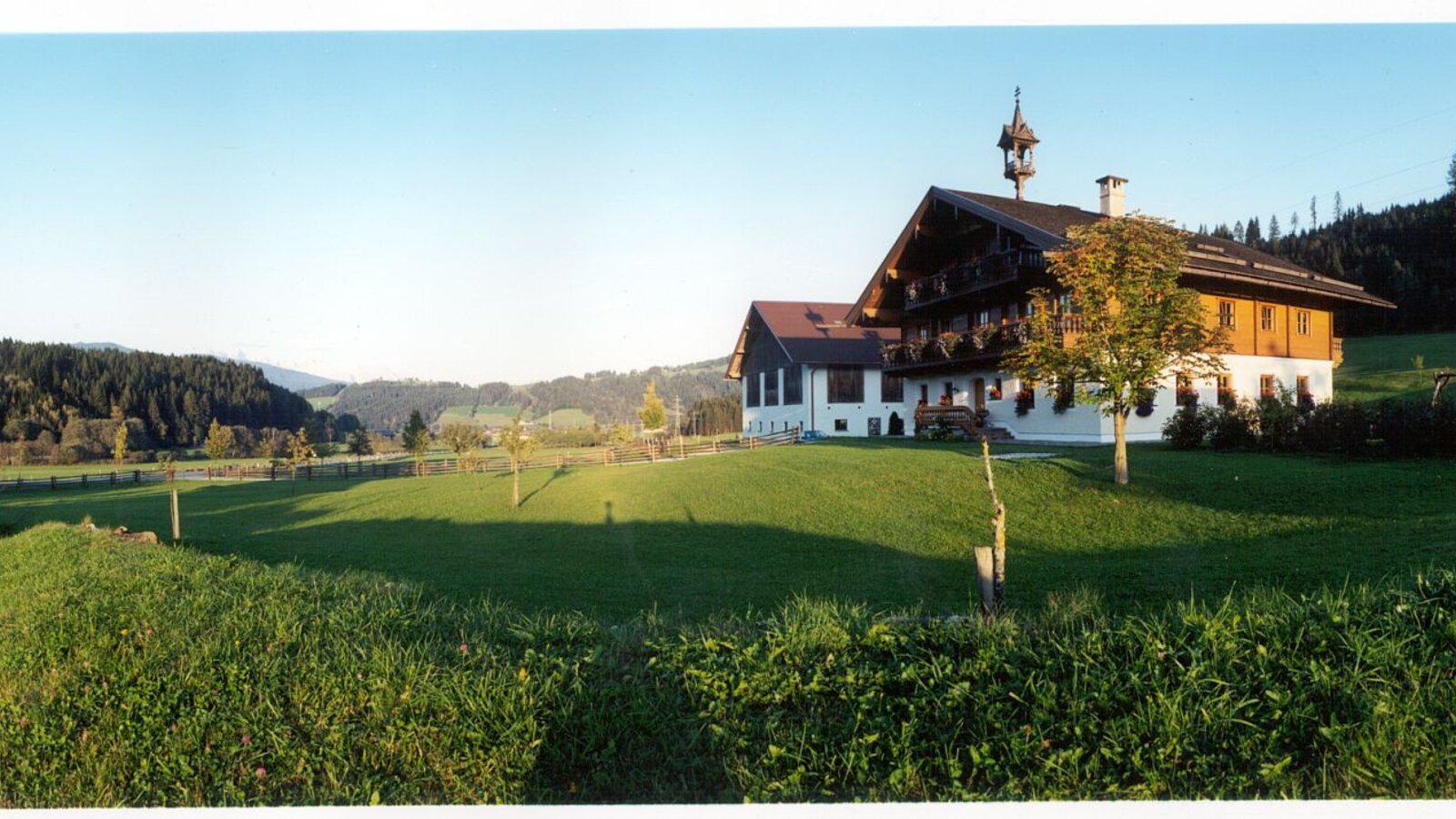 Der Bauernhof mit Glockenturm, umgeben von grünen Wiesen und bewaldeten Hügeln, inklusive Schaukel auf dem Gelände.
