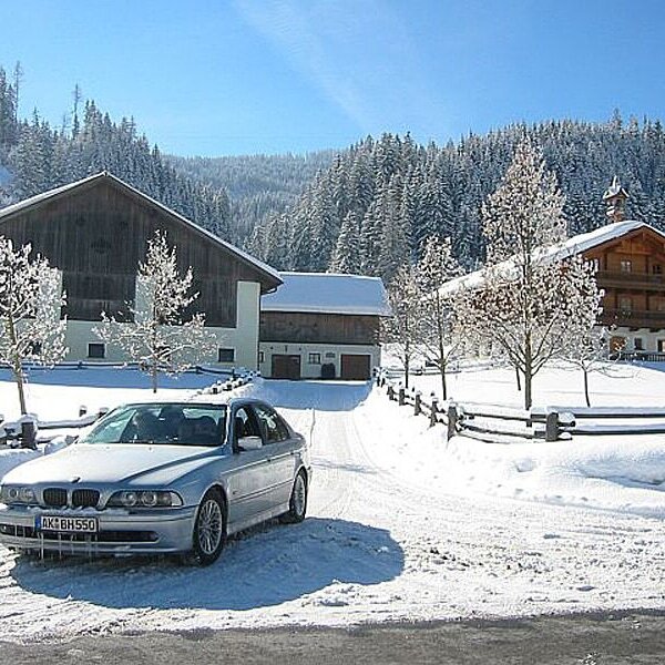 Der Bauernhof in verschneiter Winterlandschaft, mit Zufahrt und angrenzendem Wald.