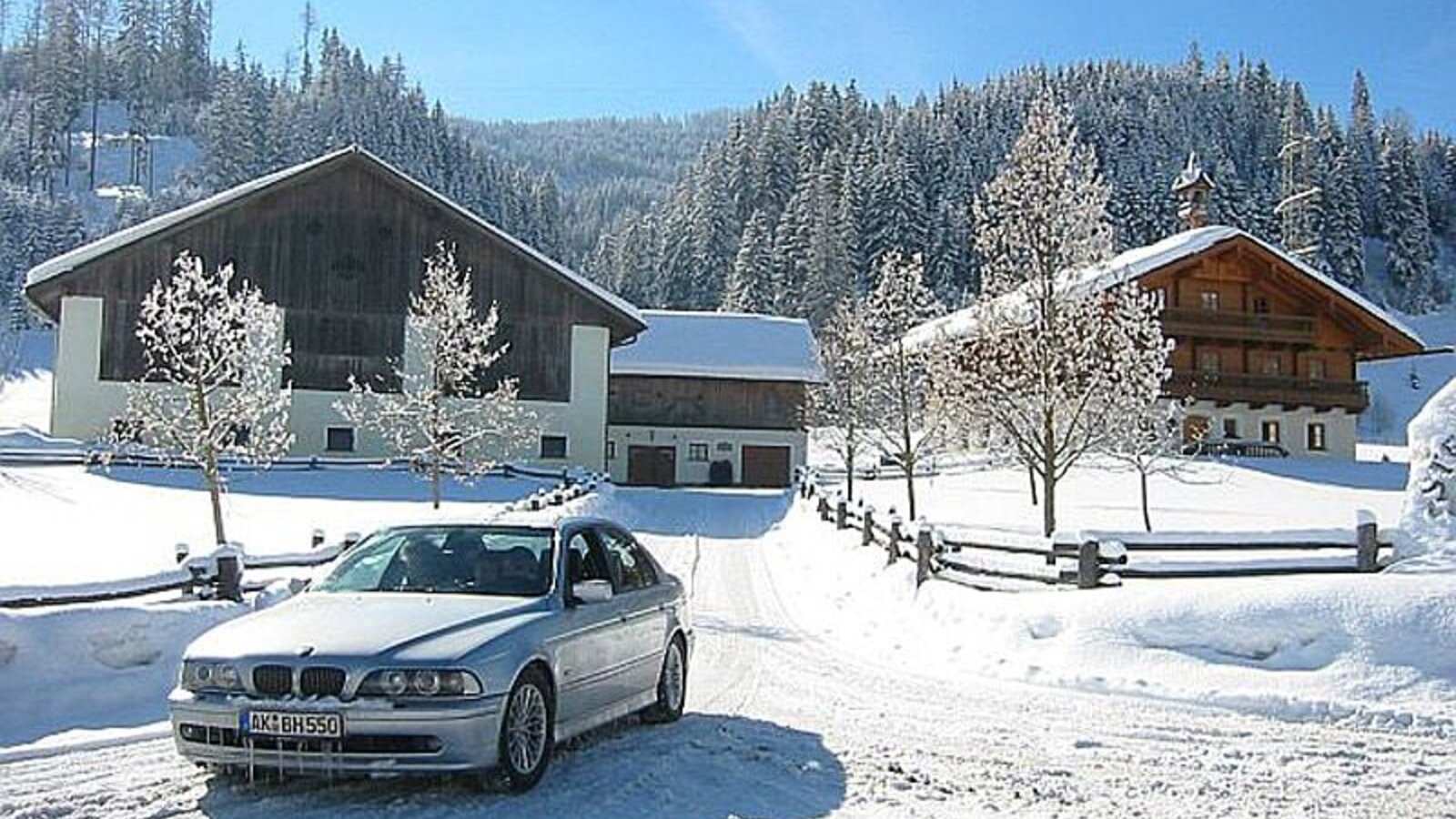 Der Bauernhof in verschneiter Winterlandschaft, mit Zufahrt und angrenzendem Wald.