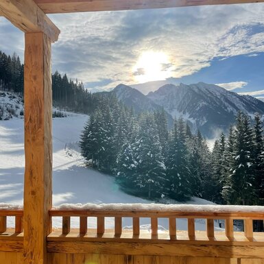 Blick vom Holzbalkon des Bauernhofs auf die verschneite Berglandschaft bei Sonnenschein.