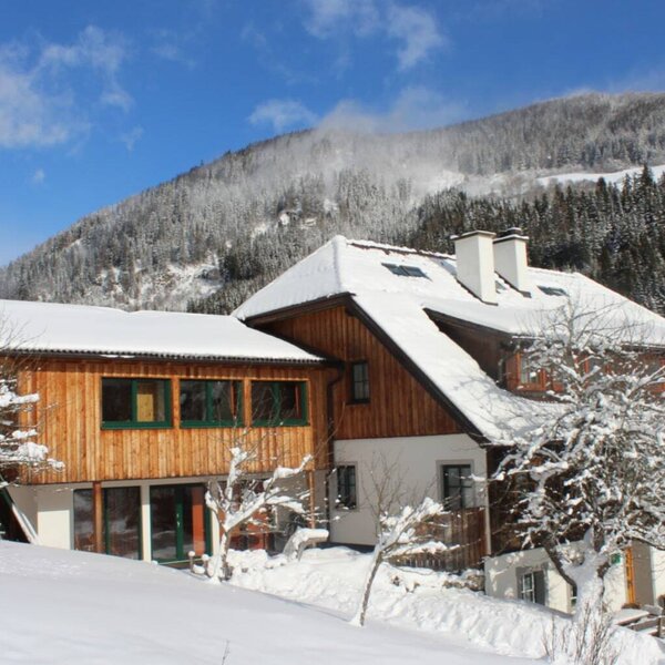 Der Bauernhof in winterlicher Umgebung mit schneebedeckten Dächern und einer Holzfassade vor der Berglandschaft.