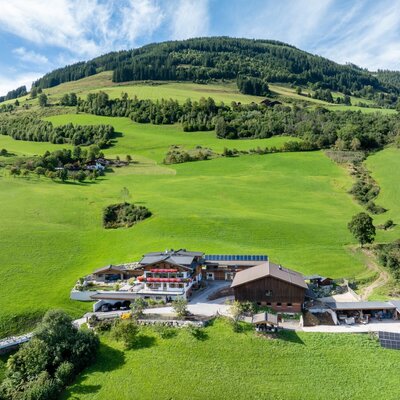 Der Bauernhof mit seinen Gebäuden, umgeben von grünen Wiesen und bewaldeten Bergen, inklusive Solaranlagen.