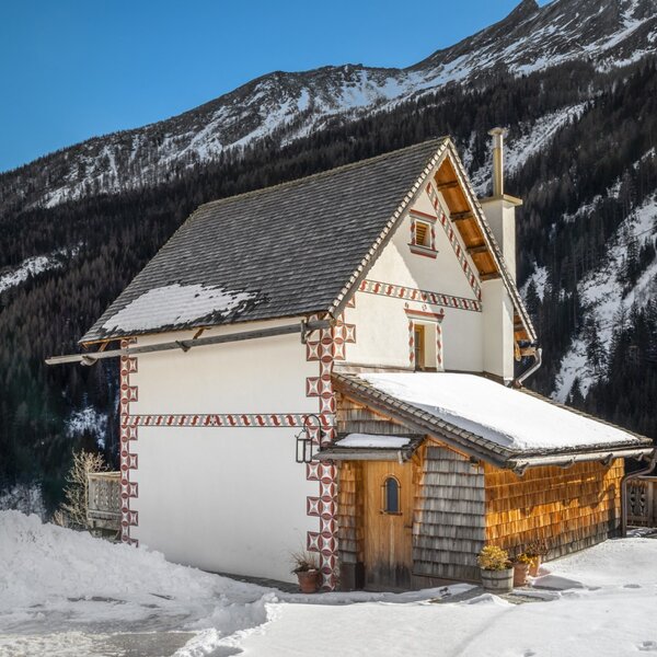 Der Hof mit traditioneller Fassadenmalerei und Schindeldach, umgeben von Schnee und Bergen.