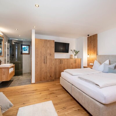 Schlafzimmer in der Ferienwohnung mit Doppelbett, Holzmöbeln, Fernseher und offenem Badbereich mit Dusche.