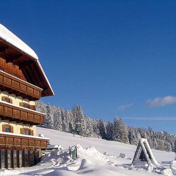 Der Bauernhof im Winter mit mehreren Balkonen und einem verschneiten Kinderspielgerät, umgeben von schneebedeckten Bäumen und Bergen.