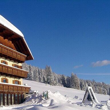 Der Bauernhof im Winter mit mehreren Balkonen und einem verschneiten Kinderspielgerät, umgeben von schneebedeckten Bäumen und Bergen.