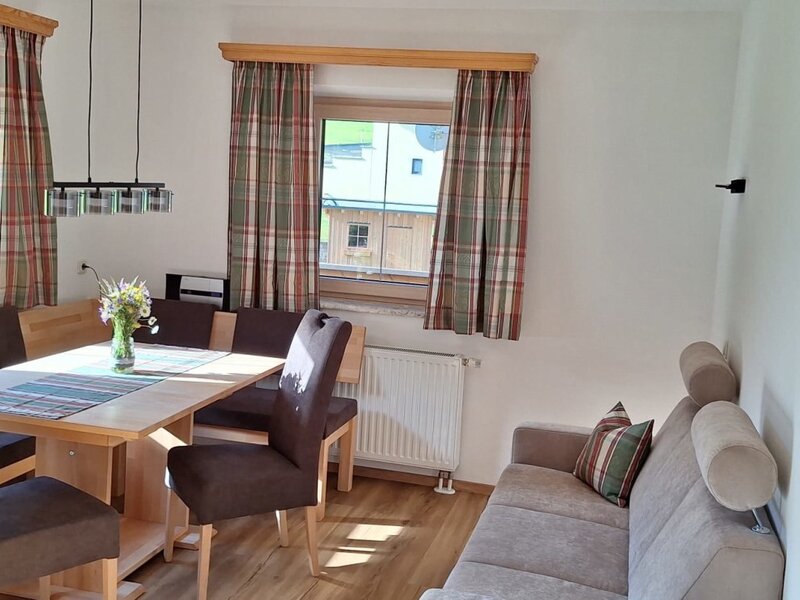Essbereich der Ferienwohnung mit Holztisch, Eckbank und Stühlen sowie einem Sofa und Fenstern mit karierten Vorhängen.