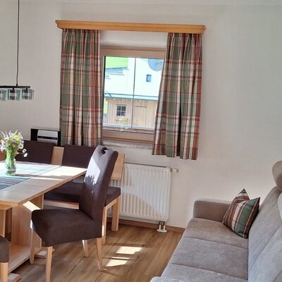 Essbereich der Ferienwohnung mit Holztisch, Eckbank und Stühlen sowie einem Sofa und Fenstern mit karierten Vorhängen.