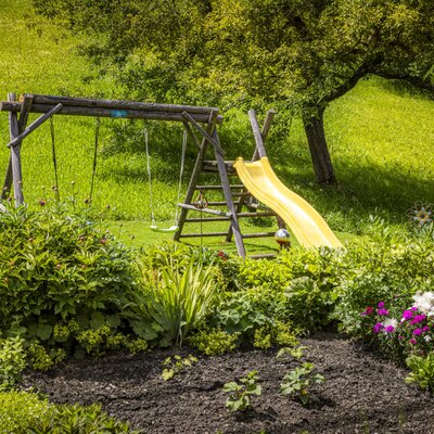 Der grüne Garten der Ferienwohnung mit einem liebevoll bepflanzten Beet und einem hölzernen Spielplatz mit Rutsche und Schaukel.
