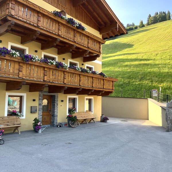 Der Bauernhof von außen mit traditionellen Holzbalkonen und Blumenkästen, einer gepflasterten Zufahrt und einem Blick auf die grünen Hügel.
