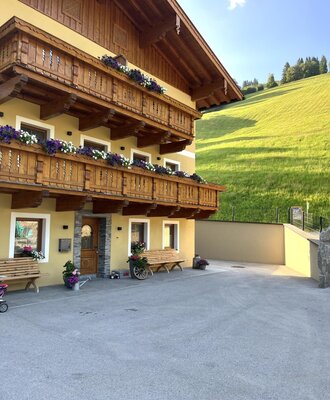 Der Bauernhof von außen mit traditionellen Holzbalkonen und Blumenkästen, einer gepflasterten Zufahrt und einem Blick auf die grünen Hügel.